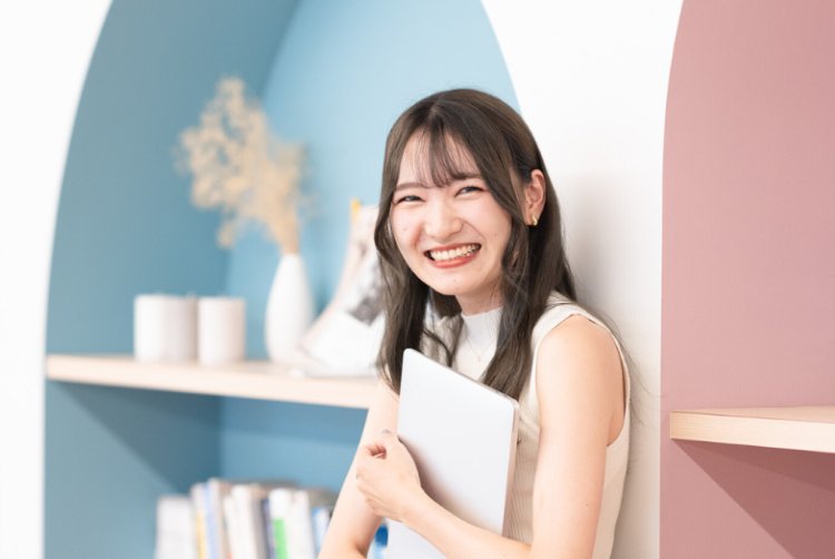 アーチ型の壁を背景にノートPCを抱え、笑顔を見せるさきさん