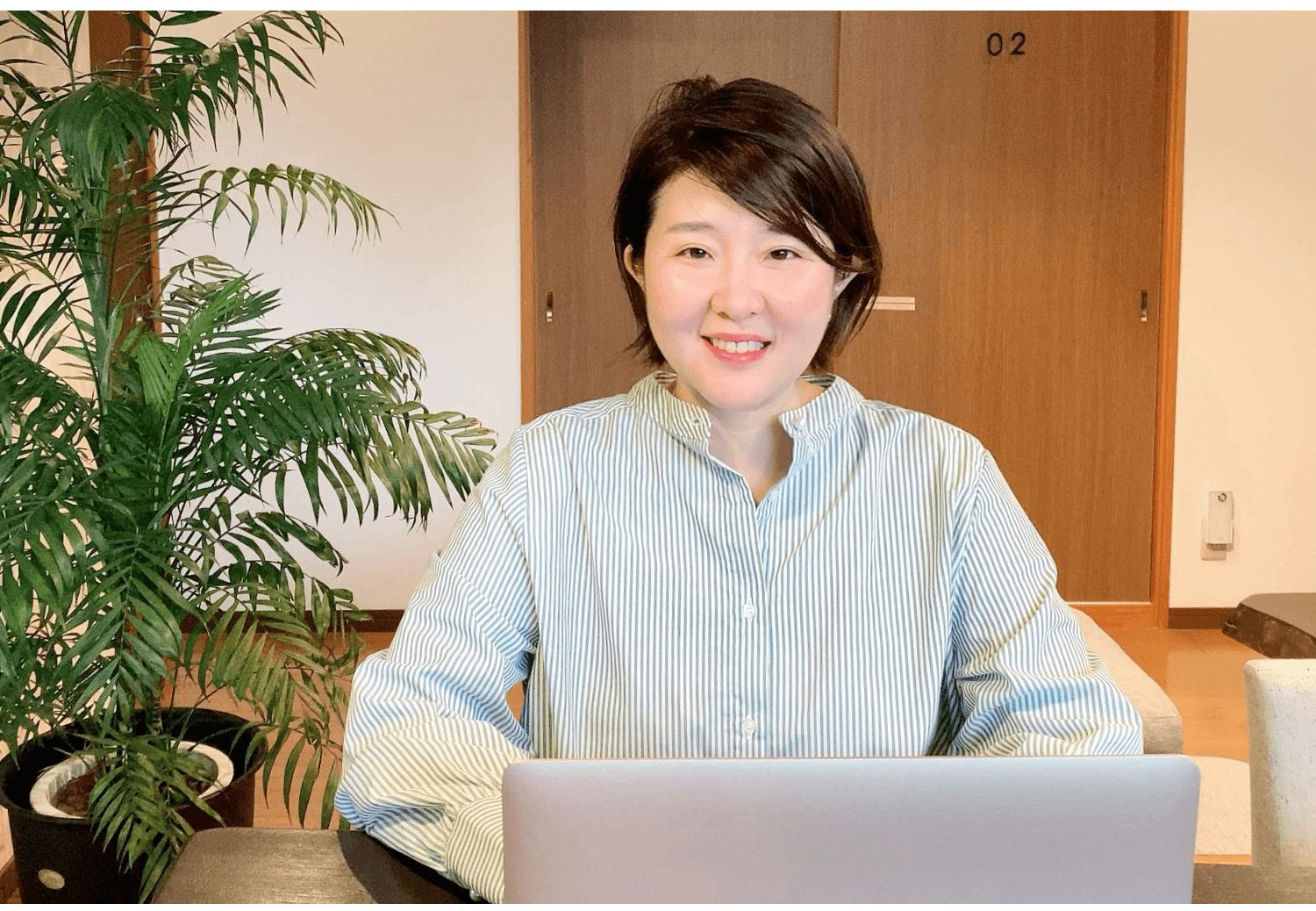 観葉植物とドア(部屋番号02)を背景に、ノートPCの前で微笑む智子さん