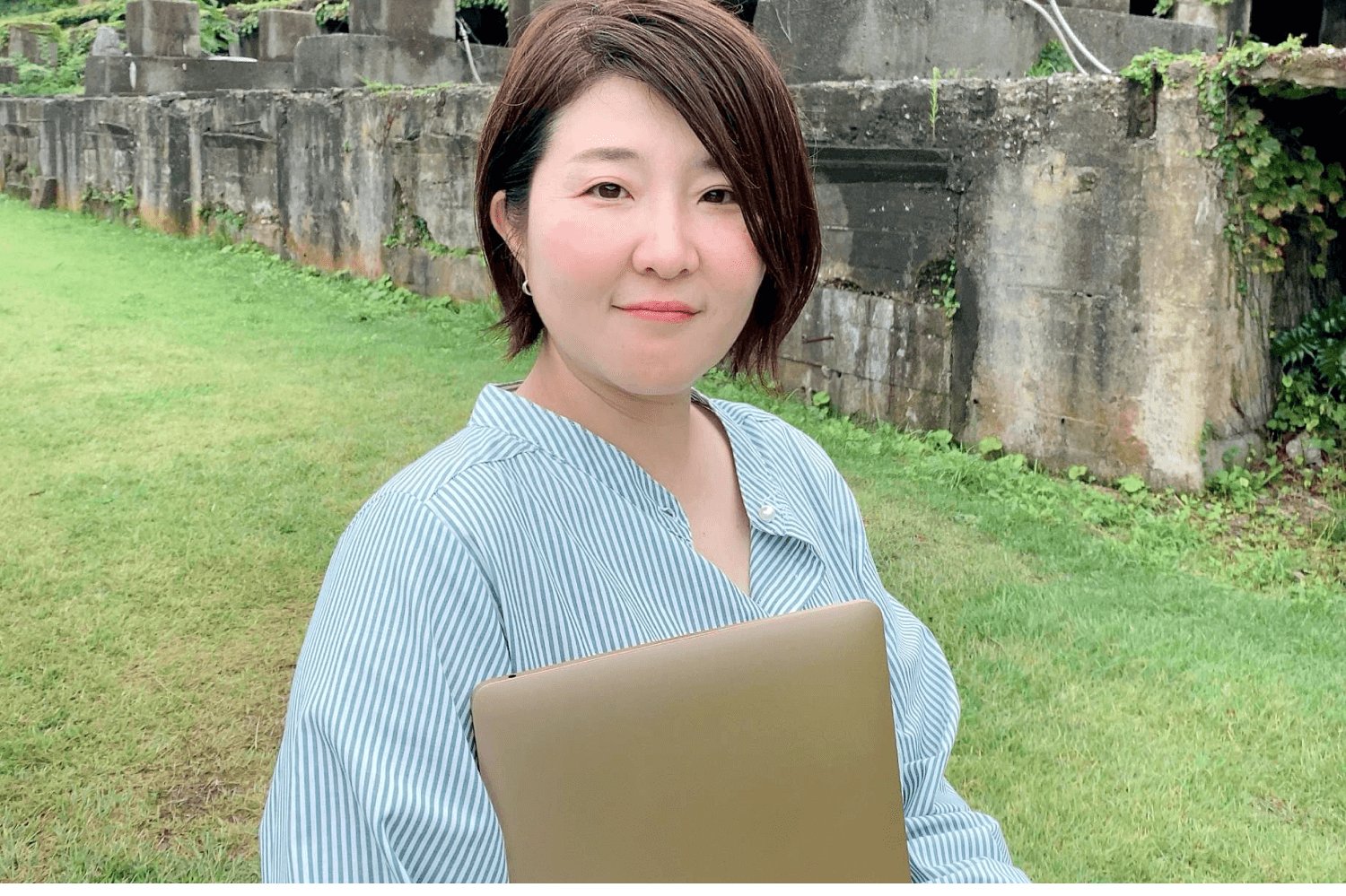 苔むした石造りの遺構を背景に、ノートPCを抱えて微笑む智子さん