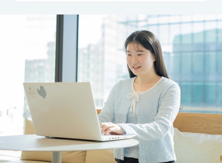オフィスの窓際でノートPCに向かい微笑むもんかさん。背景に高層ビル群