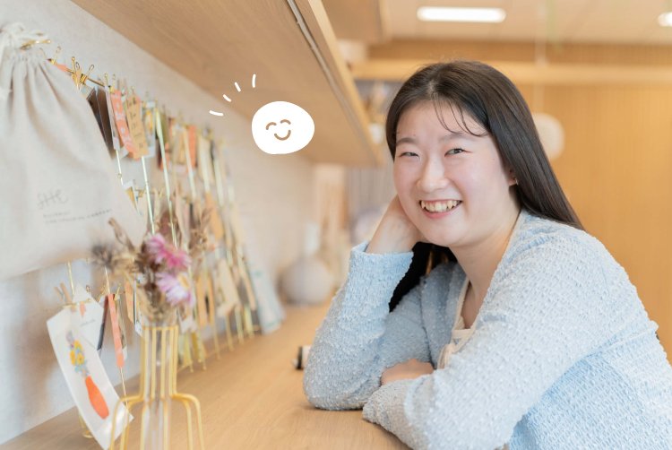 メッセージカードが並ぶ棚のそばで頬杖をつき微笑むもんかさん