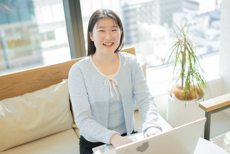 オフィスの窓際、観葉植物のそばでノートPCに向かい微笑むもんかさん