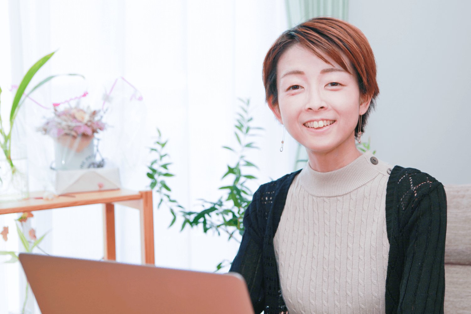 花やグリーンのある明るい部屋で、ノートパソコンを持って微笑む筆者