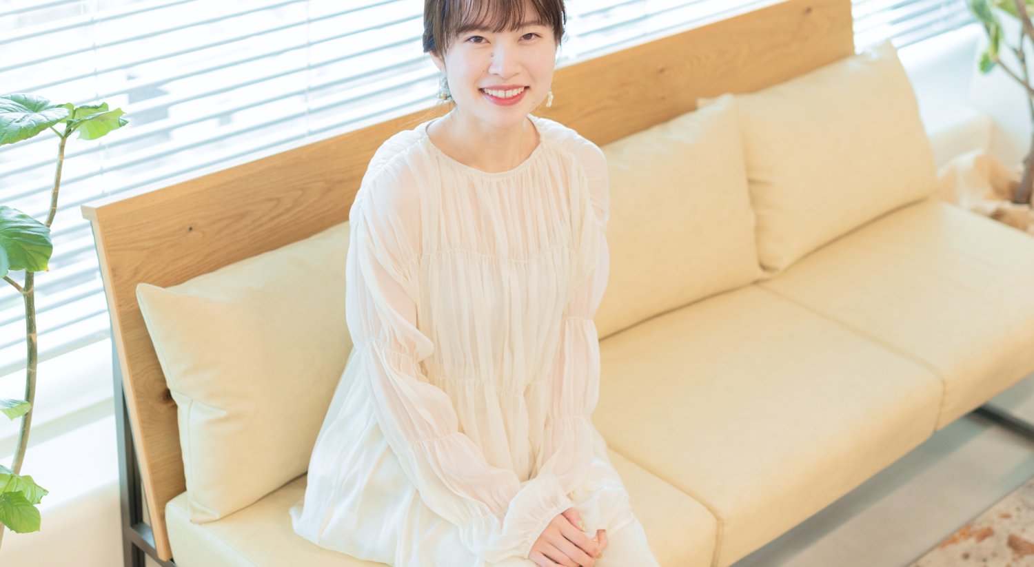 窓辺のソファに座り微笑む女性(観葉植物・ブラインド背景)