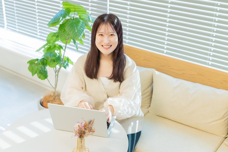 観葉植物を背景に、ソファでノートPCを操作しながら微笑むmeguさん