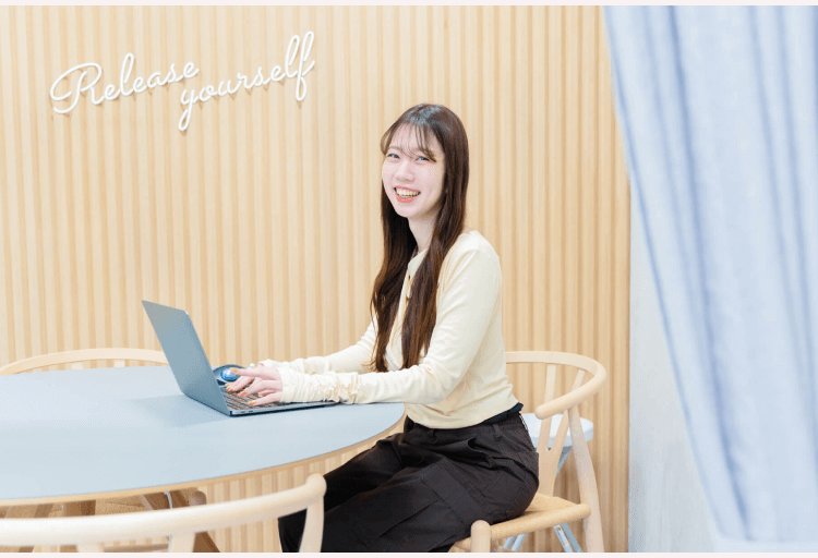 「Release yourself」の飾り文字がある部屋で、ノートPCに向かいながら振り返って微笑むしゅうまいさん