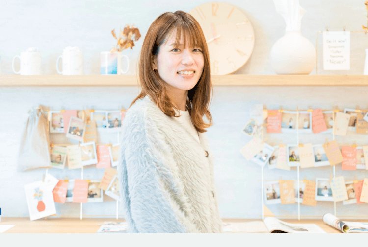 部屋の中で微笑むhisaさんの写真。棚に時計・マグカップ・写真が飾られた背景