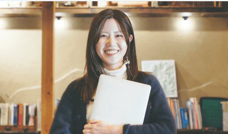 本棚を背景にノートPCを抱えて微笑むりささん