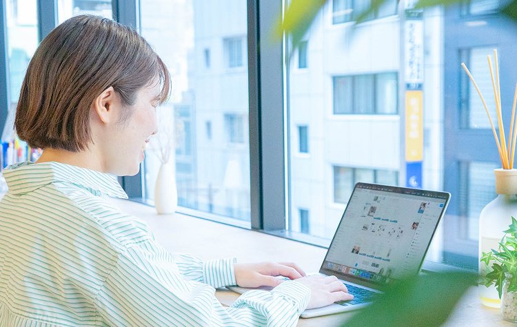 窓際のデスクでノートPCを操作するまほさんの後ろ姿
