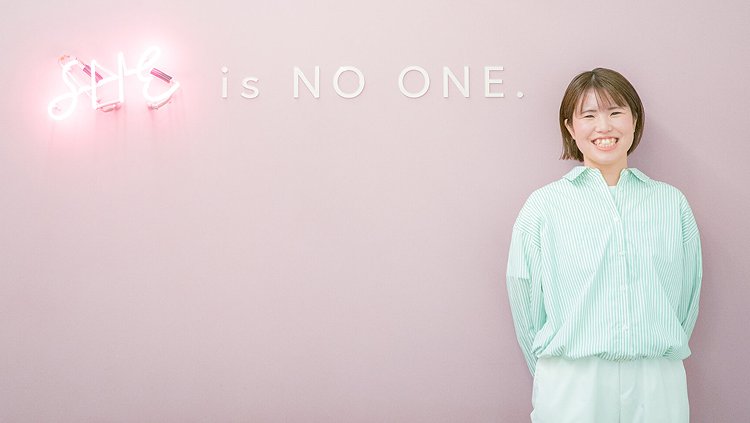 ピンクの壁に「SHE is NO ONE.」のネオンサインがある前で微笑むまほさんの写真
