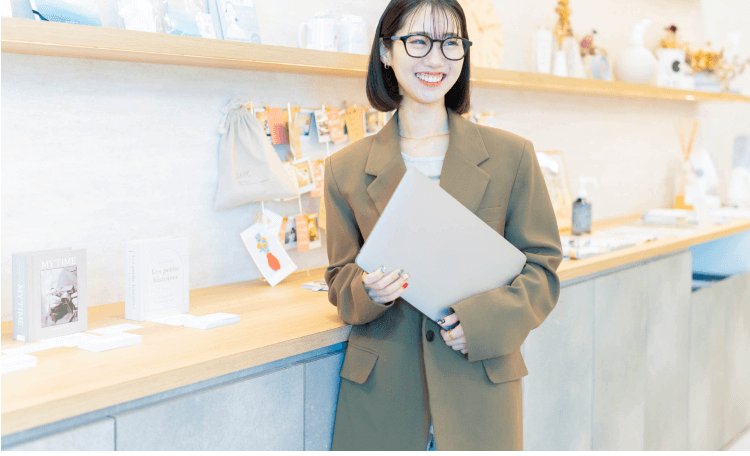 雑貨店のような店内でノートPCを抱えて微笑むまゆこさん