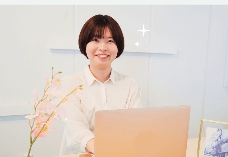 花とノートPCのある部屋で微笑む真帆さんの写真