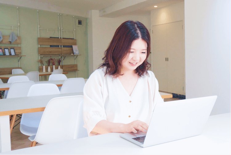 白い壁のコワーキングスペースでノートPCに向かいながら微笑むきなこさん