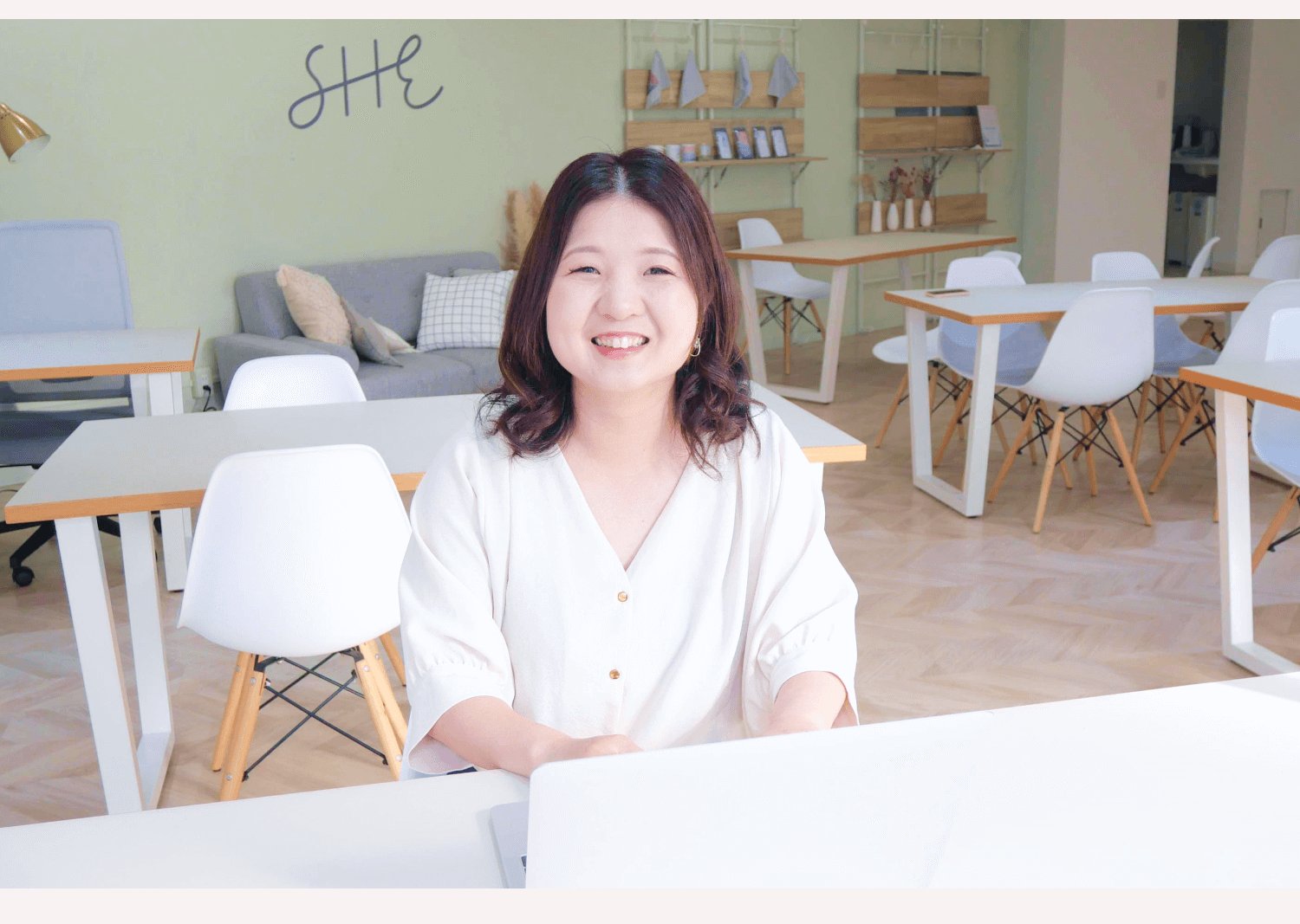 壁に「SHE」と書かれたコワーキングスペースのテーブルで微笑むきなこさん