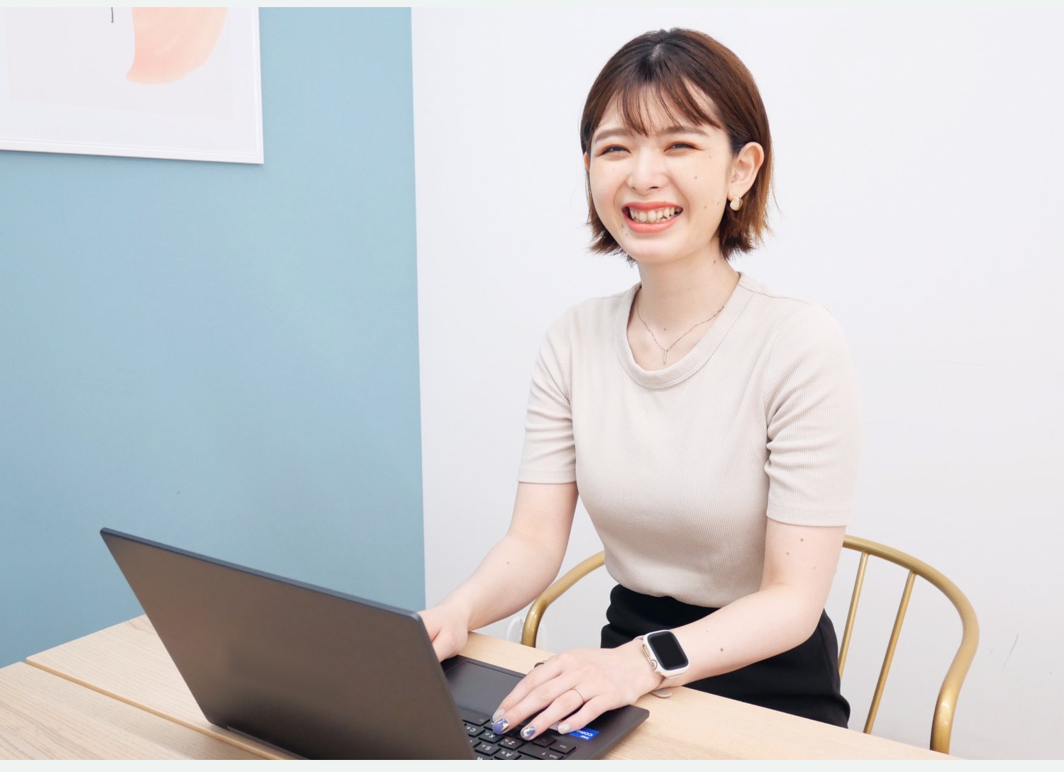 水色の壁と抽象アートを背景に、ノートPCを操作しながら笑顔を見せるシオリさん