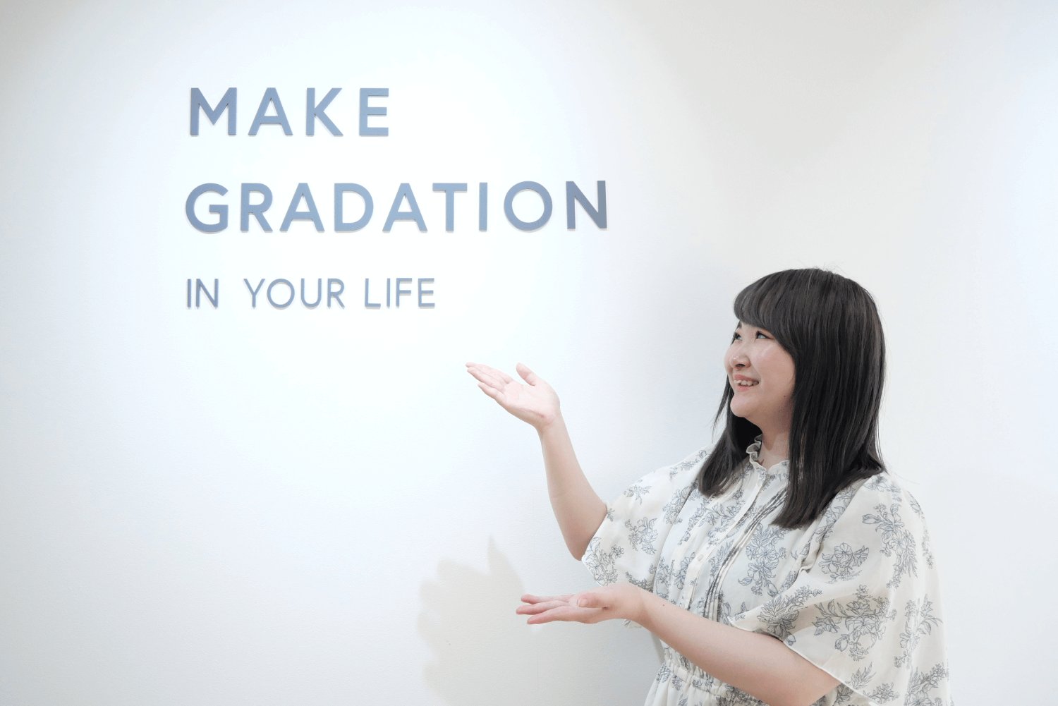 SHElikes拠点の「MAKE GRADATION IN YOUR LIFE」のロゴの前で微笑みながら手を広げるもかさん