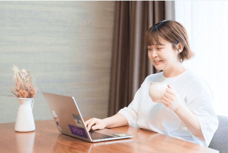 木目テーブルでドライフラワーの花瓶とノートPCを前に、マグカップを持って微笑むあかりさん