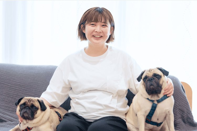 グレーのソファに座り、パグ犬2匹と一緒に微笑むあかりさん