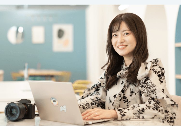 オフィスでノートPCとカメラを前に微笑むあきさんのポートレート