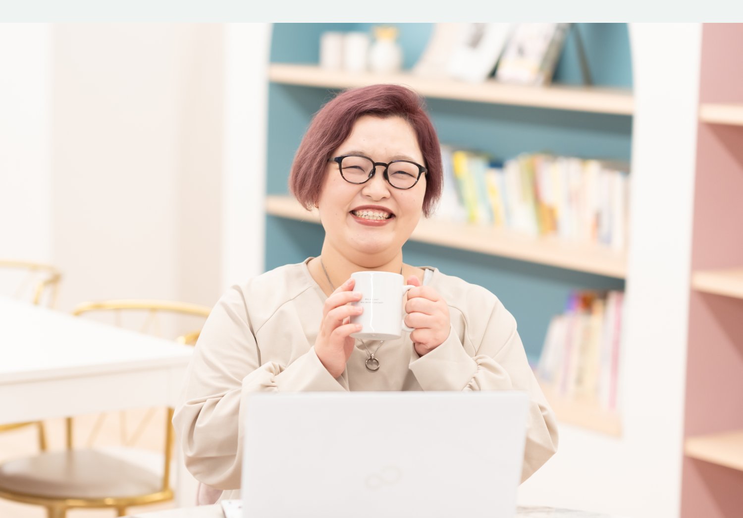 本棚を背景にマグカップを持って笑うayaさん、目の前にノートPC