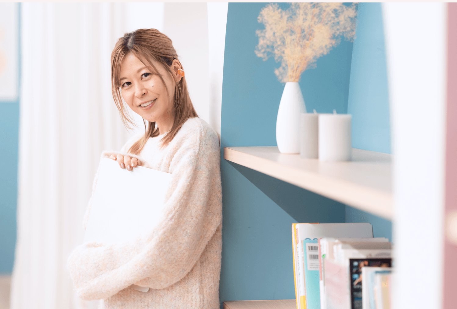 白い花瓶と青い壁を背景に、書類を抱えて微笑むゆるみさん
