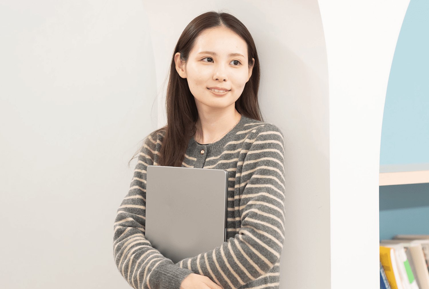白い壁にもたれ、ノートPCを胸に抱える莉央奈さん