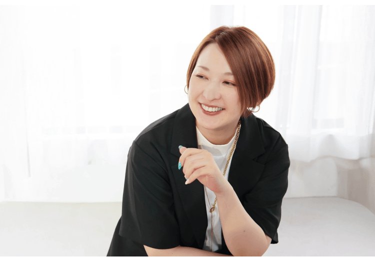 黒いシャツの女性がソファに腰掛け、ペンを手に笑顔で話しているインタビューカット